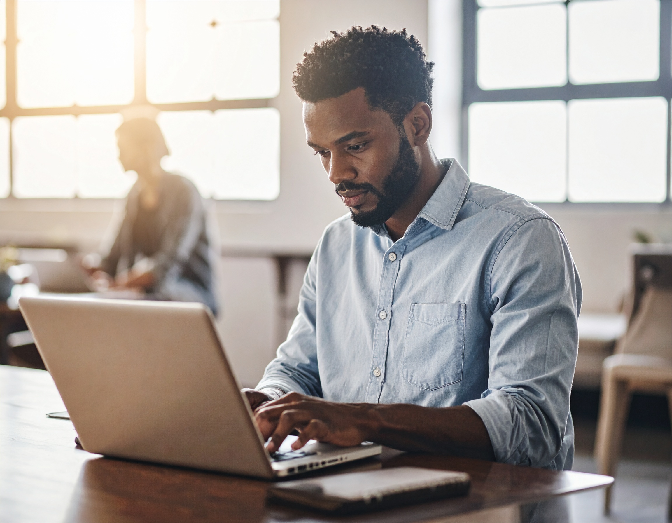 Homem trabalhando em laptop em ambiente de escritório moderno com iluminação suave de luz natural, vestido casualmente em camisa azul clara. A cena transmite concentração e profissionalismo, com fundo desfocado e janelas grandes que criam um ambiente acolhedor e iluminado.